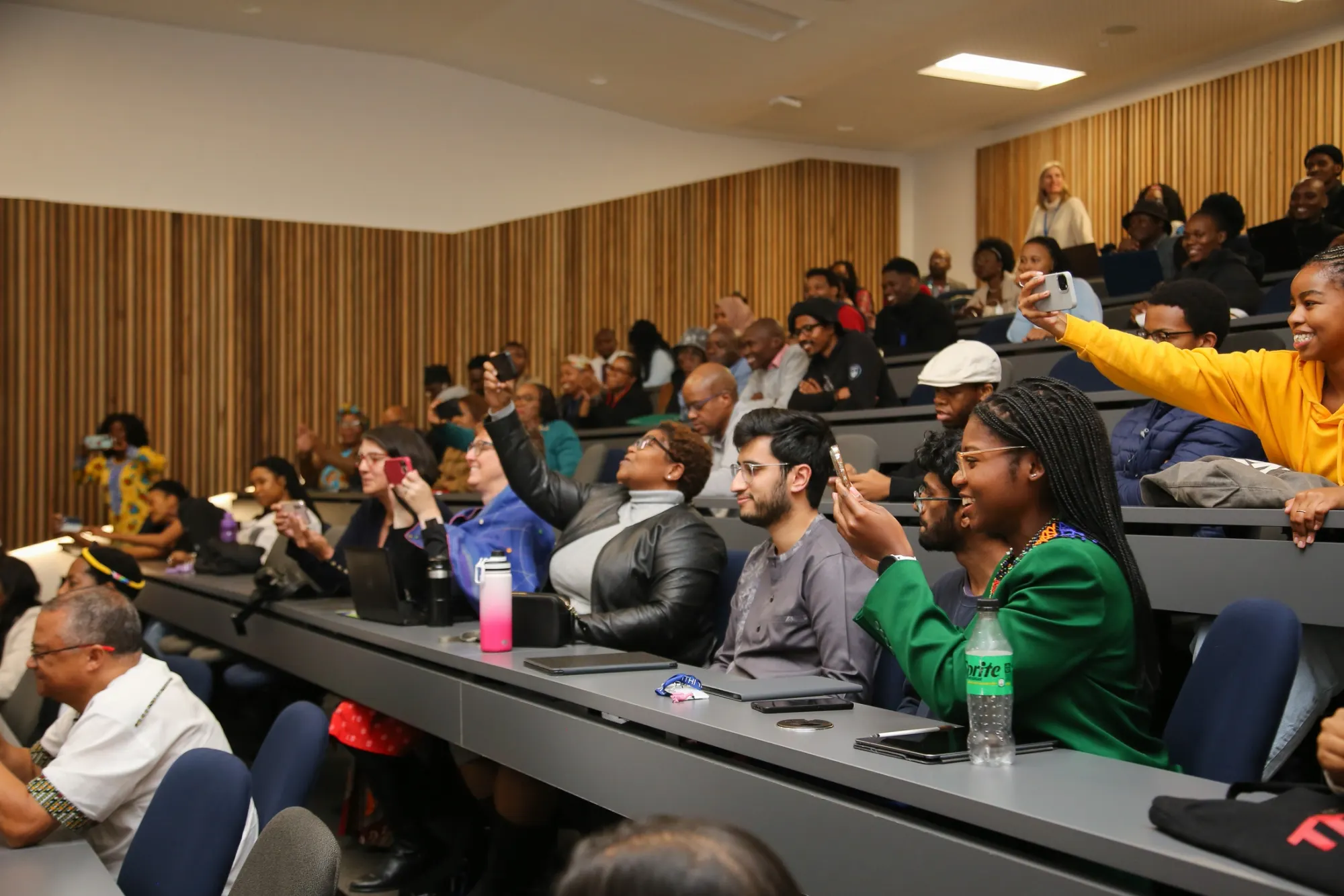 A full lecture hall of students smiling and raising phones during an ARS event